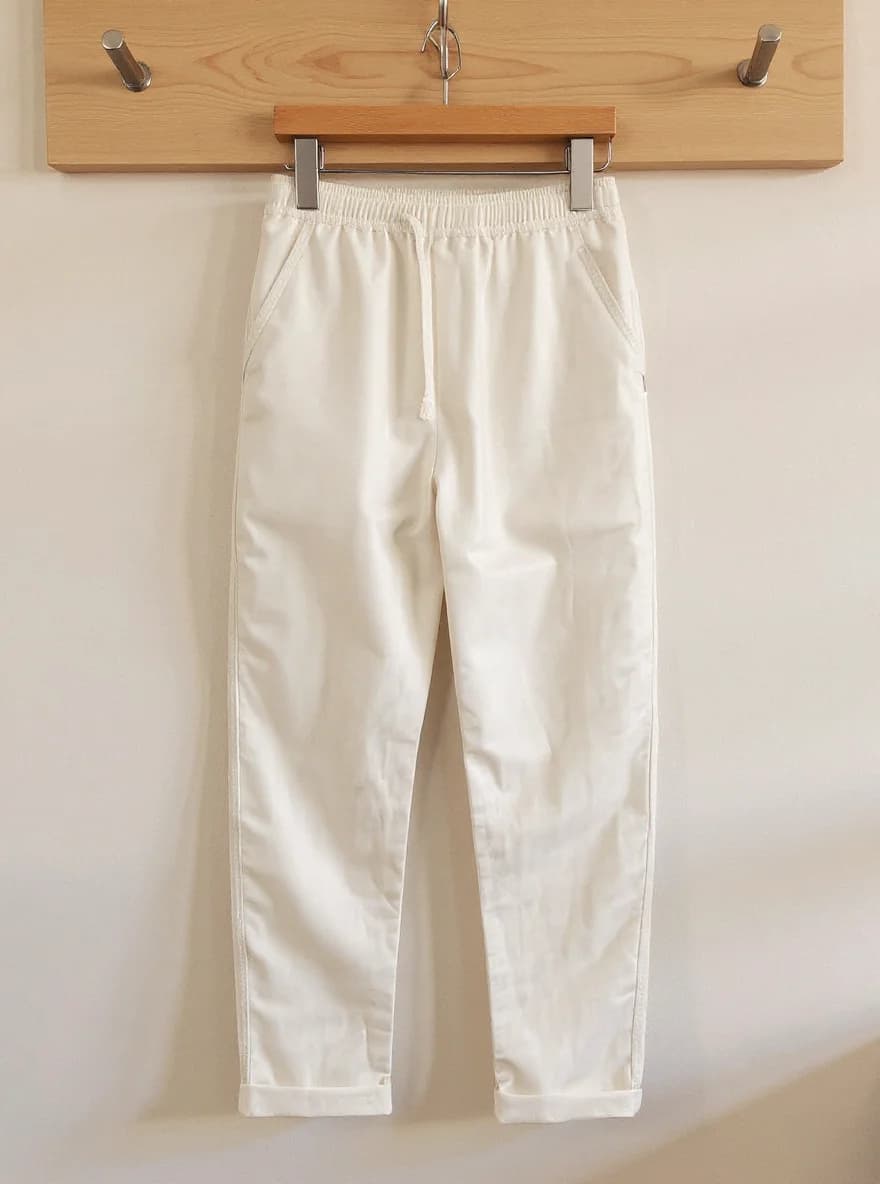 A pair of crisp white casual pants hanging on a wooden coat rack. The pants are made of a lightweight cotton material with an elastic waistband and drawstring closure. They feature side pockets and are rolled up at the ankles. The pants are photographed against a plain white wall, with the wooden coat rack mounted horizontally across the top of the frame. The coat rack has a natural wood finish and three metal hooks - two silver-colored hooks on either end and a wooden hook in the center. The pants are in pristine condition with no wrinkles or wear marks. The image is well-lit with soft, natural lighting that creates subtle shadows beneath the pants.