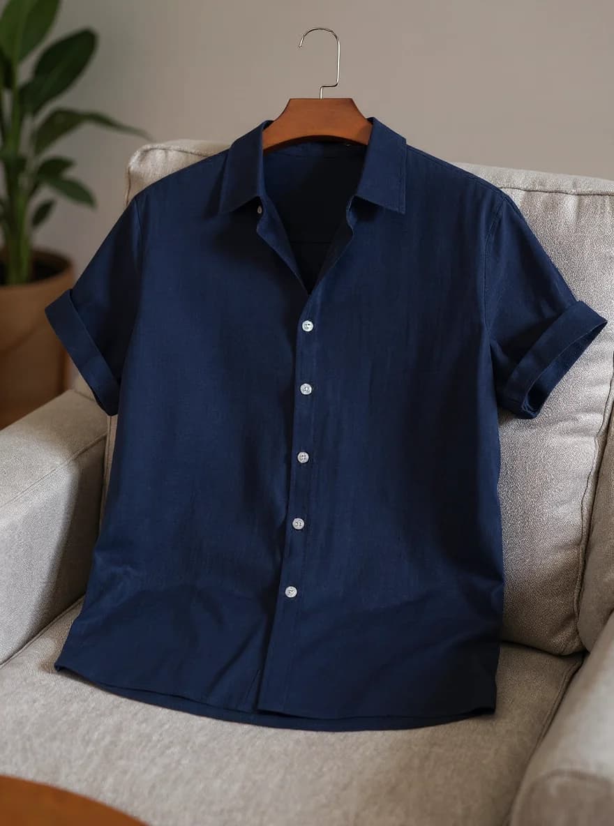 A navy blue short-sleeved button-up shirt hanging on a wooden hanger with a silver hook, displayed on a light beige upholstered armchair. The shirt has white buttons down the front and rolled-up sleeves. The fabric is a lightweight linen material with a subtle texture. The shirt is photographed against a neutral beige wall, with a green potted plant partially visible in the background. The armchair has a textured fabric upholstery and is positioned at an angle, showing its side profile. The lighting is soft and natural, creating subtle shadows beneath the shirt on the chair.