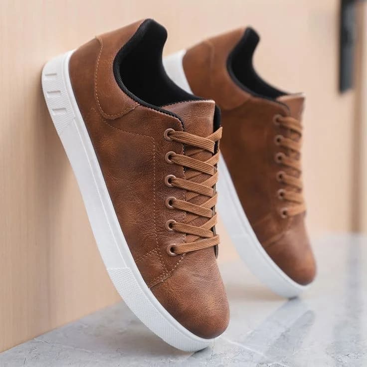 A product photo of brown leather sneakers with white rubber soles, positioned at a slight angle against a light beige wall. The shoes are photographed on a light gray concrete surface. The sneakers feature a classic low-top design with brown laces threaded through metal eyelets. The leather upper has a distressed, vintage texture with subtle variations in the brown coloring. The shoes have black lining visible at the collar and tongue. The white sole has a chunky, modern design with a slight platform effect. The lighting is soft and even, creating subtle shadows beneath the shoes. The image is cropped to show just the front and side profile of the pair, with the right shoe slightly behind the left, creating depth in the composition.