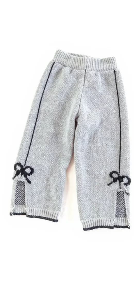 The clothing item is a pair of knitted pants or joggers. They are primarily light gray with black accents. The pants feature a ribbed, elastic waistband and ribbing at the cuffs. There are two vertical black lines running down the sides of each leg. Additionally, there are black bow designs with a checkered pattern on the lower sides of each leg, near the cuffs. The material appears to be soft, possibly a knitted or woolen fabric.. A pair of light gray knitted children's pants photographed against a white background. The pants feature a ribbed elastic waistband and are cropped in length. The bottom hem and side panels have decorative black bows with white centers. The pants have a textured knit pattern throughout and a diagonal seam running down the front. The cuffs at the bottom are ribbed with a striped pattern in black and white. The pants are photographed laying flat and centered in the frame, with natural shadows cast beneath them. The image has a clean, minimalist composition with the pants as the sole focus against the stark white backdrop.