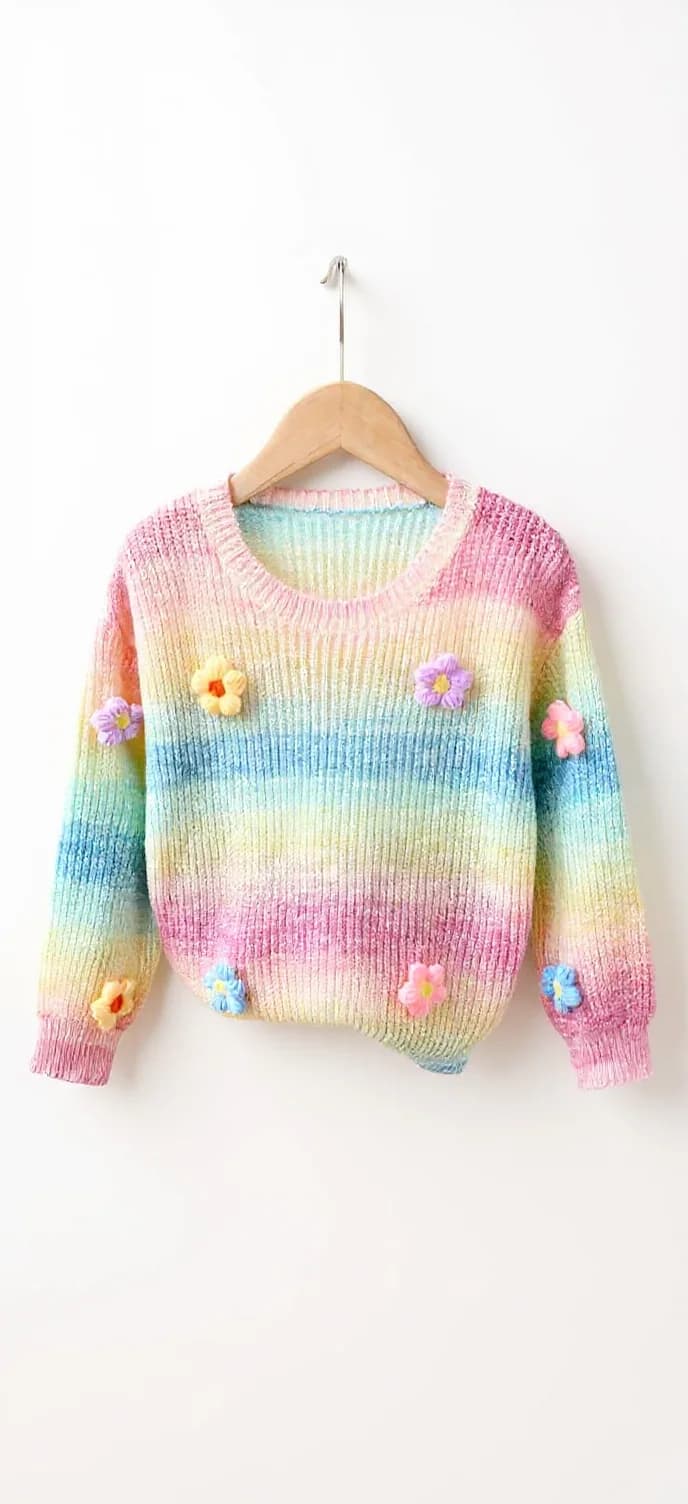 This is a knitted sweater with horizontal pastel rainbow stripes in pink, yellow, green, blue, and purple. It features a ribbed crew neckline and long sleeves with ribbed cuffs. The sweater is decorated with small, three-dimensional flower embellishments in various pastel colors, positioned on the front and sleeves. The material appears soft and textured, typical of knitted fabric.. A pastel rainbow-colored knit sweater hanging on a wooden hanger against a plain white background. The sweater features horizontal stripes in soft colors including blue, yellow, pink, and green. Small floral appliques in yellow, pink, and blue are scattered across the front of the sweater. The sweater has a crew neckline, ribbed cuffs, and a cropped length. The knit pattern is a classic ribbed texture throughout. The sweater is photographed straight-on, centered in the frame, with the wooden hanger's hook visible at the top. The image has a clean, minimalist composition with ample white space around the garment.