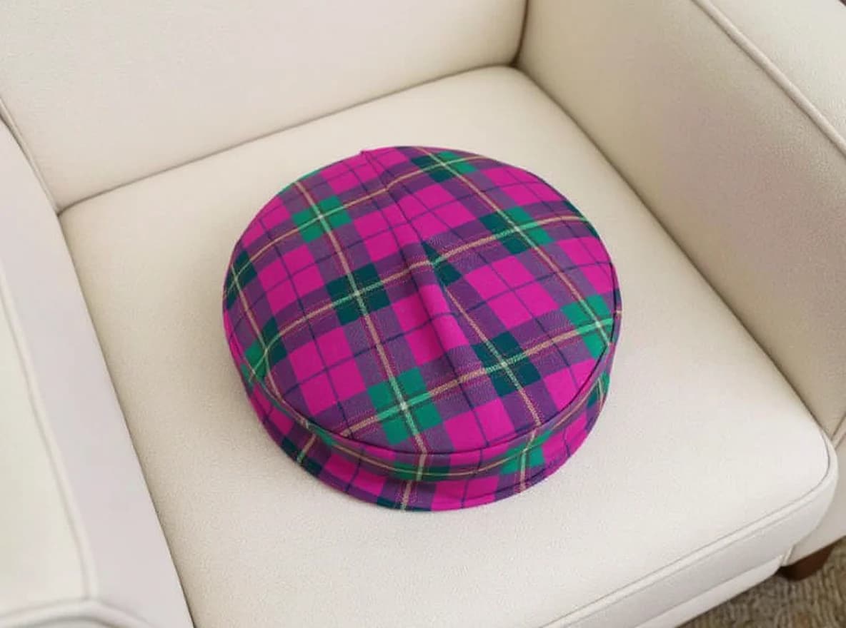 A round, plaid fabric cushion or of pillow with a vibrant pattern. The pattern includes shades of pink, green, yellow, and purple in a tartan design. The fabric appears to be soft and woven, suitable for use as a cushion or decorative pillow.. A bright pink and green tartan plaid round ottoman cushion placed on a white leather armchair. The ottoman cushion features a traditional Scottish plaid pattern with hot pink as the primary color, intersected by forest green and navy blue lines creating a classic checkered design. The cushion is perfectly circular and has a slight indentation in the center, showing it is soft and pliable. The white leather chair is partially visible, showing clean lines and a modern, minimalist style. The image is shot from a slightly elevated angle, capturing the full circular shape of the ottoman against the stark white background of the chair.