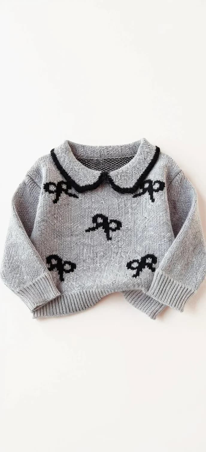 This is a knitted sweater in light gray color. It features a pattern of black bows scattered across the fabric. The sweater has a round neckline with a gray collar edged in black. The sleeves are long, with ribbed cuffs, and the hem is also ribbed. The material appears to be soft and textured, typical of knitwear.. A gray knit sweater with a Peter Pan collar and black trim. The sweater features a repeating pattern of black bows scattered across the front. The bow design is simple and stylized, with each bow having a curved shape and a small tail. The sweater has long sleeves and is photographed laid flat against a white background. The knit texture of the sweater is clearly visible, showing a ribbed pattern. The collar has a pointed shape typical of a Peter Pan collar, and the black trim runs along the neckline, shoulders, and sleeve cuffs. The sweater is cropped to show only the upper portion, with the bottom hem partially out of frame.