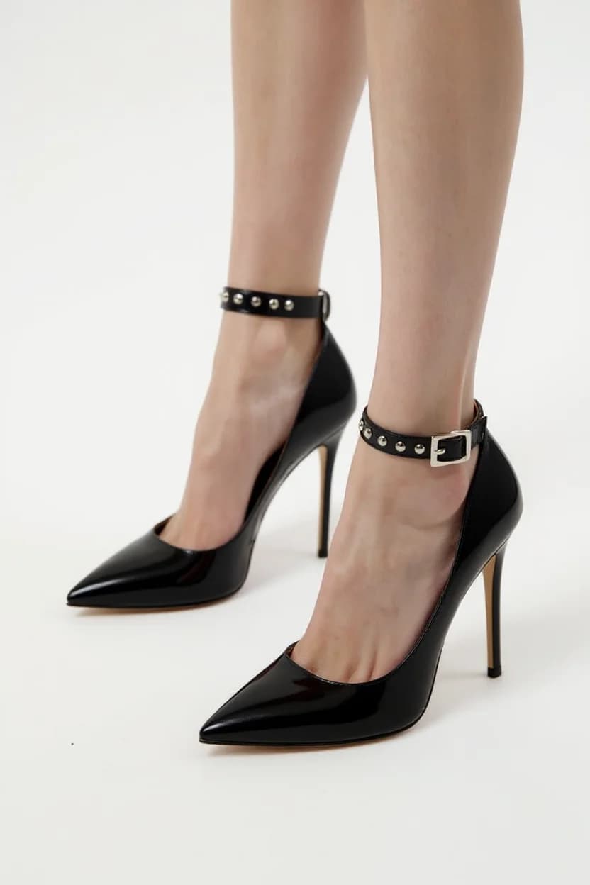 A product photography shot of black patent leather high heel shoes with an ankle strap detail. The shoes feature a pointed toe, stiletto heel 4 inches tall, and a sleek ankle strap decorated with silver metal studs and a small rectangular buckle. The shoes are worn on a person's feet, showing them against a pure white background. The image is cropped to focus on the lower legs and feet, with the shoes positioned at a slight angle to show both the side and front profiles. The lighting is bright and even, creating a clean, professional studio look. The shoes have a glossy finish that reflects light, emphasizing their luxurious appearance.