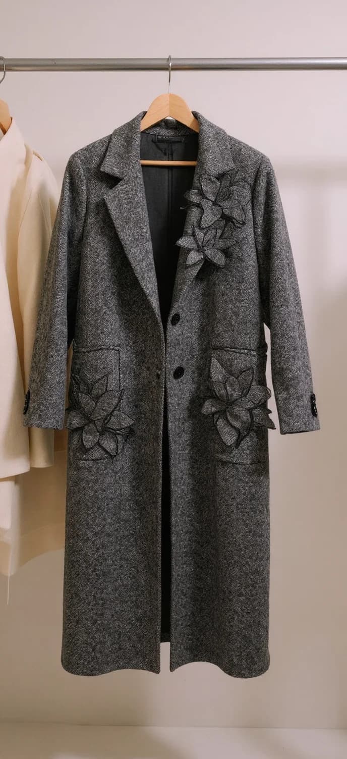 This is a long, gray, woolen coat with a textured pattern. It features large, embroidered floral appliqués on the front, with three on the right side and two on the left. The coat has a classic lapel collar, long sleeves with buttoned cuffs, and two large front pockets. It is designed with a straight fit and has button closures down the center.. A high-quality product photo of a gray wool coat hanging on a wooden hanger against a plain white wall. The coat is a long-length style with distinctive flower-shaped appliques on both front pockets. The coat features a notched lapel collar, black buttons down the front, and a tailored silhouette. The wool fabric has a subtle heathered texture throughout. The coat is photographed in a vertical format, showing the full length from shoulders to hem. A partial view of another garment in cream color is visible on the left edge of the frame. The lighting is soft and even, creating subtle shadows that highlight the texture of the wool and the dimensional quality of the floral appliques.