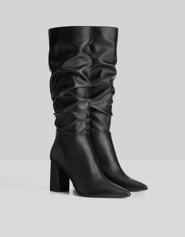 A product photo of a pair of black leather knee-high boots with a ruched design. The boots feature vertical gathers across the upper portion, creating a slouchy, elegant silhouette. The boots have a pointed toe and a chunky block heel 3 inches tall. The leather has a smooth, matte finish and drapes elegantly from top to bottom. The boots are photographed against a plain white background, positioned at a slight angle to show both the side profile and front details. The lighting is even and professional, casting subtle shadows beneath the boots. The image shows only the boots, with no other accessories or styling elements in frame.