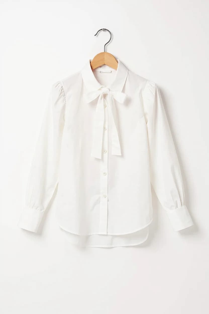 A crisp white long-sleeved blouse hanging on a wooden hanger against a plain white background. The blouse features a classic pointed collar, a front bow tie detail at the neck, and a button-down front with small white buttons. The sleeves are gathered at the wrists with a slight puff at the shoulders. The fabric is a lightweight cotton material with a smooth, matte finish. The blouse is photographed straight-on, centered in the frame, with soft, even lighting that eliminates harsh shadows. The wooden hanger has a natural finish and a silver metal hook. The image has a minimalist, clean aesthetic with ample negative space around the garment.