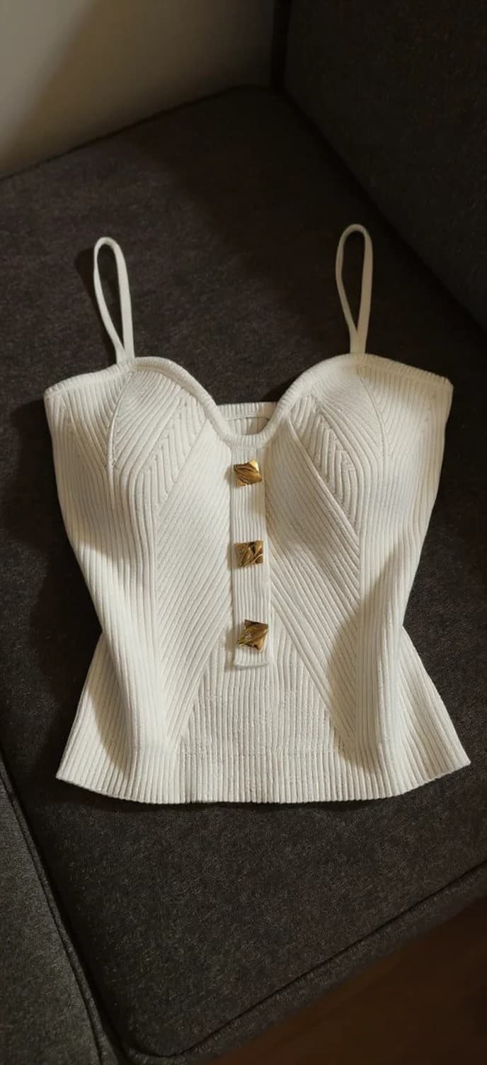 A high-quality product photograph of a white ribbed crop top on a dark gray textured fabric background. The top features a corset-style design with three gold-toned decorative buttons down the center front. The fabric has a diagonal ribbed texture pattern throughout. The garment has thin white straps and a peplum-style waist that creates a fitted silhouette. The top is photographed from a straight-on angle, slightly elevated, casting subtle shadows on the dark background. The lighting is soft and directional, highlighting the texture of the white fabric while creating gentle shadows that emphasize the garment's structured design.