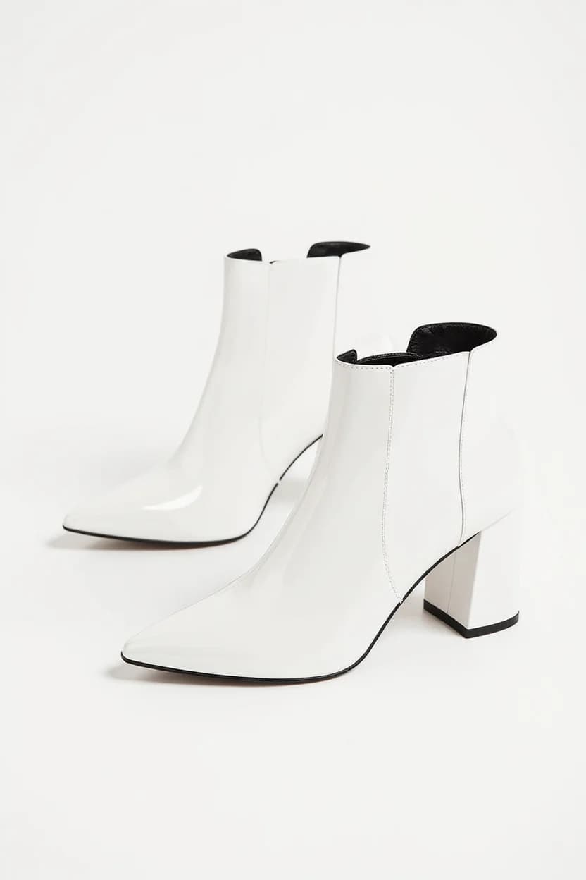 A product photo of white leather ankle boots with a chunky block heel against a pure white background. The boots have a pointed toe design and a Chelsea boot silhouette with elastic side panels. The heel is 3 inches tall and has a geometric, angular shape in white. The boots are photographed from a side angle, showing both boots placed at a slight diagonal. The leather has a smooth, polished finish that creates subtle shadows. The interior lining of the boots is black, visible at the opening. The boots cast a soft shadow beneath them on the white surface. The image has a clean, minimalist aesthetic with sharp detail and high contrast between the white boots and background.