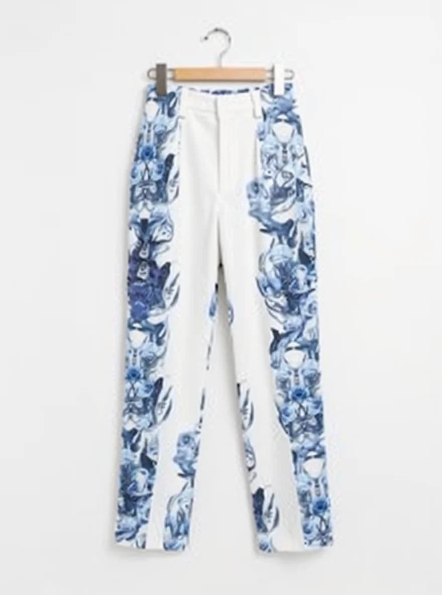 A pair of white dress pants with a blue floral print pattern hanging on a wooden hanger against a white background. The pants feature a distinctive blue watercolor-style print of roses and leaves scattered across the fabric. The print is concentrated more densely on the sides and lower portion of the pants, creating a graduated effect. The pants have a straight-leg cut, high waist, and are made of a lightweight fabric suitable for formal wear. The wooden hanger has a natural brown finish and silver metal hook. The pants are photographed in a straight-on, product-style shot with clean lighting that highlights the contrast between the white base fabric and the blue floral design.