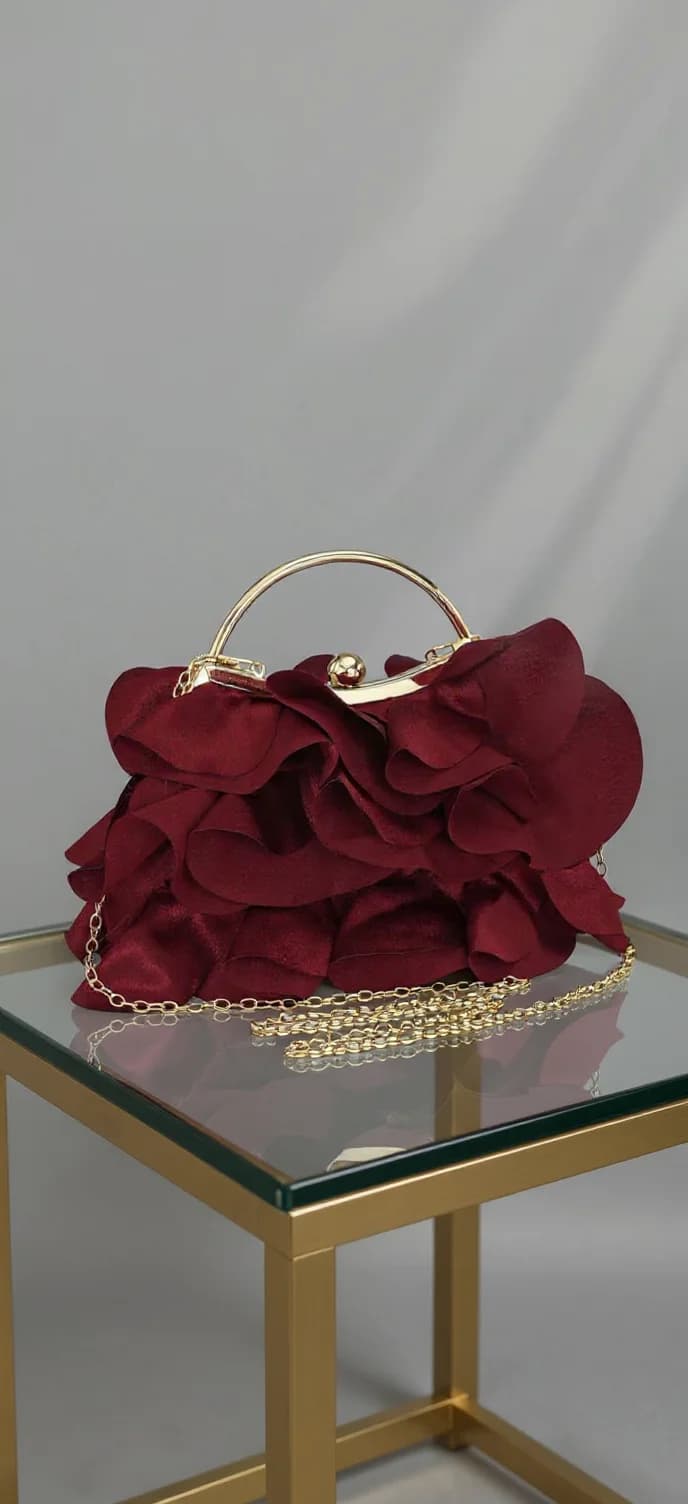 A luxurious burgundy evening clutch purse photographed on a glass and gold metal side table against a plain light gray background. The purse features a dramatic ruffled design with multiple layers of deep burgundy fabric petals creating a flower-like pattern. The purse has a curved gold metal frame handle and is secured with a gold clasp. A delicate gold chain strap is attached to the sides of the purse. The glass table top has a reflective surface that creates a subtle mirror effect beneath the purse. The lighting in the image creates soft shadows on the gray backdrop, and the composition is centered with the purse taking up the majority of the frame. The purse's fabric has a rich, velvet-like texture that catches the light, highlighting the dimensional quality of the ruffles.