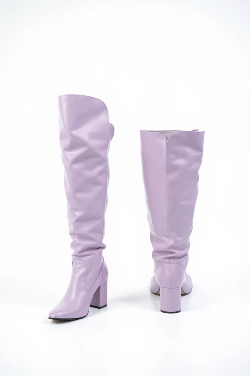 A product photo of a pair of light lavender knee-high boots against a pure white background. The boots are made of smooth leather with a slouchy, relaxed fit design. The boots feature a chunky mid-height heel 3 inches tall and a pointed toe. The left boot is positioned at a three-quarter angle showing the side profile, while the right boot is turned to show the back and heel detail. The boots have a relaxed, gathered silhouette from the knee down, creating soft pleats in the leather. The lighting is bright and even, casting subtle shadows beneath the boots. The image has a clean, minimalist aesthetic with sharp focus and high resolution detail showing the texture of the leather material.