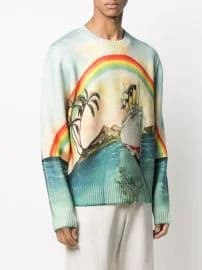 A product photo of a light blue sweater with a whimsical tropical island scene. The sweater features a large rainbow arching across the chest area, with vibrant red, orange, yellow, green, and blue bands. The rainbow connects to a small island with palm trees on the left side and a lighthouse on the right side. The island is rendered in a watercolor-style illustration with turquoise and mint green tones. The sweater has a crew neckline and is made of a knit material. The model is wearing white pants and the photo is taken against a plain white background. The sweater has a relaxed, oversized fit with dropped shoulders and ribbed cuffs. The artwork on the sweater is painted in a soft, artistic style with visible brush strokes and a dreamy, ethereal quality.