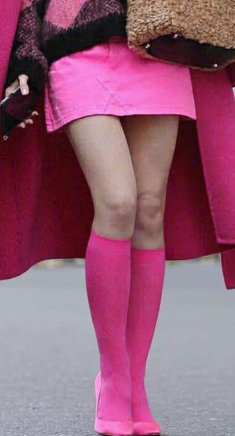 A fashion-focused photograph showing a person's lower body wearing bright pink knee-high socks and a pink mini skirt. The image is cropped to focus on the legs and skirt, with the top portion of the outfit partially visible. The person is wearing a black and pink plaid sweater, and a brown fur-trimmed coat is visible at the top edge of the frame. The background is a light gray concrete surface. The lighting is even and natural, highlighting the vibrant pink color scheme of the outfit. The image is a close-up shot that emphasizes the bold color coordination between the pink skirt, socks, and sweater.