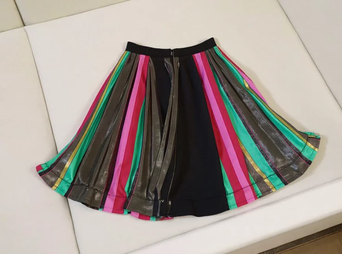 The product is a pleated skirt featuring vertical stripes in various colors, including pink, green, black, yellow, and metallic shades. The skirt has a wide, black waistband and includes a center zipper with a metallic pull. The pleats are evenly spaced, giving a structured and voluminous appearance to the skirt. The material appears to be a shiny, possibly satin or similar fabric, with a smooth and reflective texture.. A pleated midi-length skirt displayed on a white surface. The skirt features a black base fabric with vertical stripes in multiple colors - mint green, hot pink, and metallic gold accents. The stripes are arranged in a fan-like pattern, creating a dramatic A-line silhouette. The skirt has a black elastic waistband and a silver zipper closure running down the center back. The pleats are crisp and evenly spaced, creating a flowing, elegant appearance. The skirt is photographed from above at a slight angle, showing the full length of the garment. The white surface beneath the skirt is a table, and the image is well-lit with no harsh shadows.