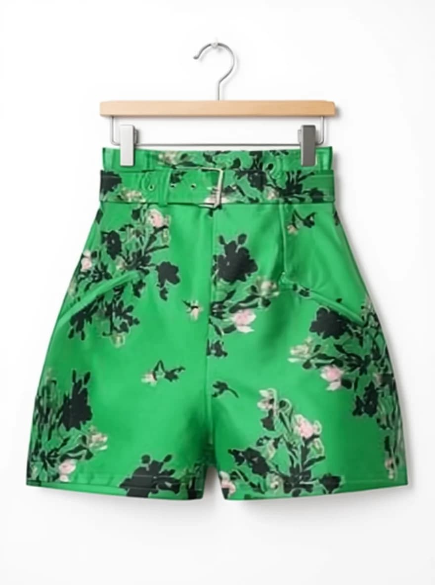 A pair of high-waisted green shorts hanging on a wooden hanger against a white background. The shorts feature a bold floral pattern with black and pink flowers scattered across the bright kelly green fabric. The shorts have a fitted waistband with a belt loop detail, and a paperbag-style waist. The hem falls at mid-thigh length. The shorts are made from a structured fabric with a slight sheen. The wooden hanger has a natural finish and a silver metal hook at the top. The shorts are photographed straight-on, creating a clean product shot style.