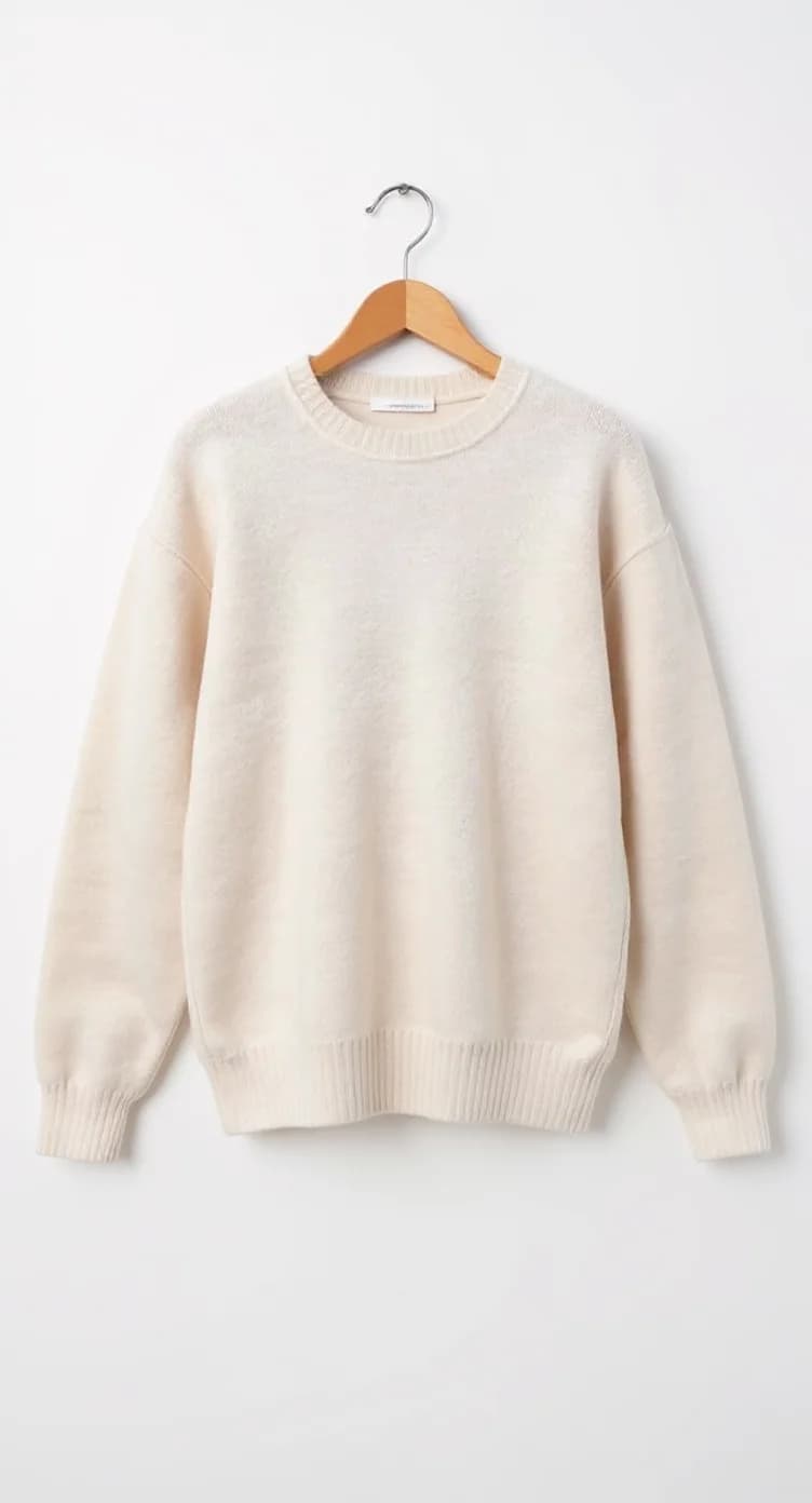 A cream-colored crew neck sweater hanging on a wooden hanger against a plain white background. The sweater has a relaxed, oversized fit with dropped shoulders and long sleeves. The ribbed hem and cuffs are visible at the bottom and sleeves. The sweater is made of a soft knit material with a matte finish. The wooden hanger has a silver metal hook at the top and a natural wood finish. The sweater is photographed straight-on, creating a clean, minimalist product shot. The image has soft, even lighting that eliminates harsh shadows, creating a professional studio-quality photograph.