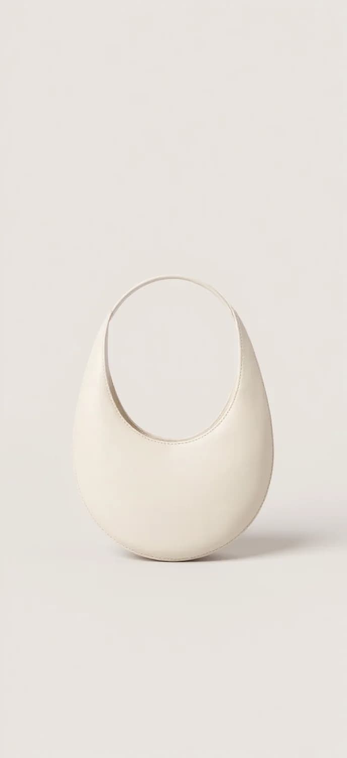 A minimalist, sculptural ceramic bowl photographed against a plain white background. The bowl has a smooth, curved form with a deep, bowl-like depression in the center. The piece is made of white porcelain with a matte finish. The bowl's shape is asymmetrical, with one side being taller than the other, creating an organic, flowing silhouette. The lighting is soft and even, casting subtle shadows beneath the piece. The bowl is positioned slightly off-center in the frame, photographed from a straight-on perspective. The surface texture is uniform and unmarked, with clean lines and precise craftsmanship. The overall composition has a modern, artistic quality with strong emphasis on form and negative space.