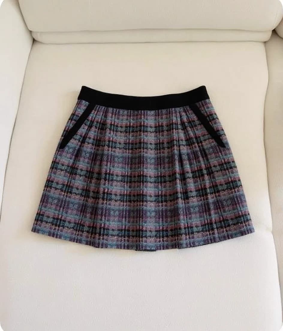 A plaid wool skirt photographed on a white leather couch. The skirt features a black elastic waistband and is pleated throughout. The plaid pattern consists of blue, pink, and gray colors creating a classic tartan design. The skirt has a mini-length cut that falls above the knee. Two black fabric pockets are visible at the hip level. The skirt is photographed from a straight-on angle, centered on the light beige leather couch. The image has a clean, minimalist composition with the skirt as the sole focus against the neutral background.
