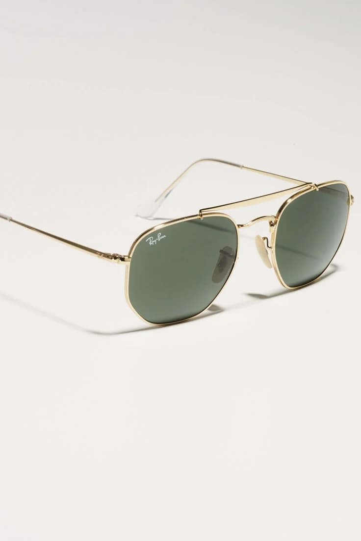 A product photo of Ray-Ban sunglasses on a plain white background. The sunglasses have a gold-colored metal frame with a double bridge design and aviator-style shape. The lenses are dark green with a slight gradient tint. The frame has thin metal temples extending backward. The Ray-Ban logo is visible on the left lens in a small, subtle engraving. The sunglasses are positioned at a slight angle, with the right temple arm partially out of frame. The lighting is soft and even, creating subtle shadows beneath the sunglasses. The image has a clean, professional product photography style with sharp focus and high detail resolution.